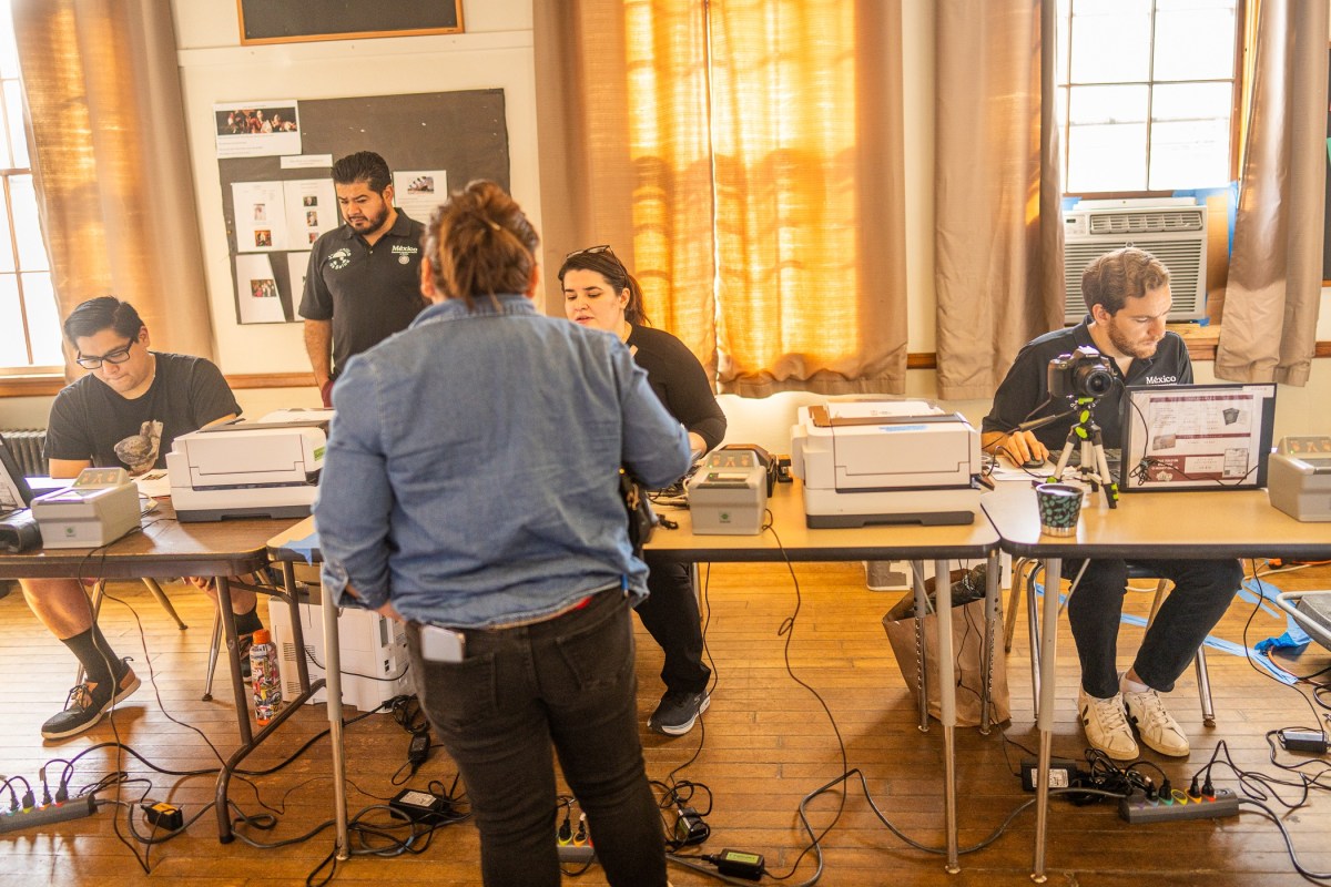 Five people work at two tables with computers, printers, and various equipment in a room with wooden floors and large windows with tan curtains.