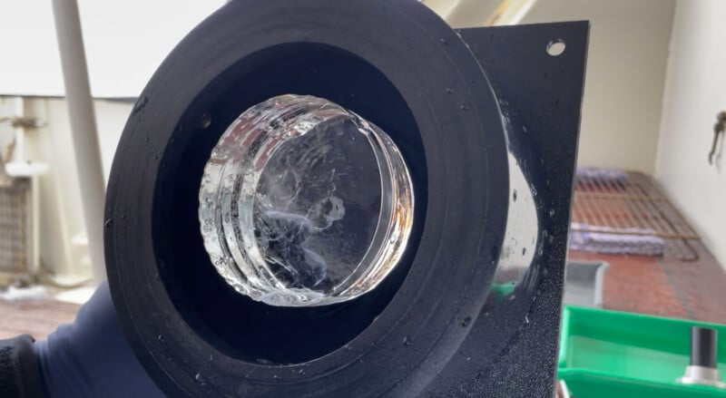 Close-up of a thick, circular layer of clear ice forming inside a round black metal frame, with water droplets on the surface and blurred background elements.