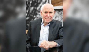 An older man with short white hair, wearing a dark suit jacket and light blue shirt, sits at a table and looks at the camera with a gentle expression. The background is blurred and patterned.