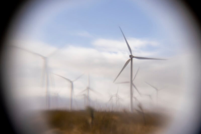 A wind farm with several wind turbines stands on grassy land under a partly cloudy sky, viewed through a blurry, circular frame that gives a hazy, vignette effect to the image.