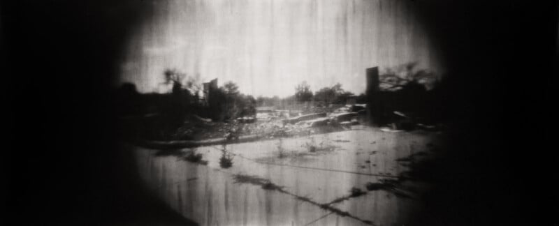 A black and white, blurry, vignetted image of an abandoned, overgrown lot with scattered debris, leafless trees, and distant structures under a cloudy sky.