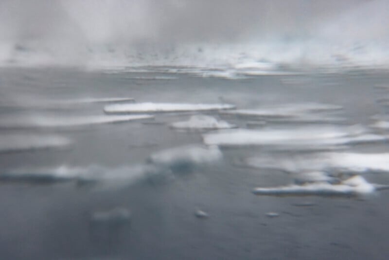 Blurry photo of ice floes floating on a cold, gray body of water, with mist or fog obscuring the background. The overall scene appears cold and desolate.