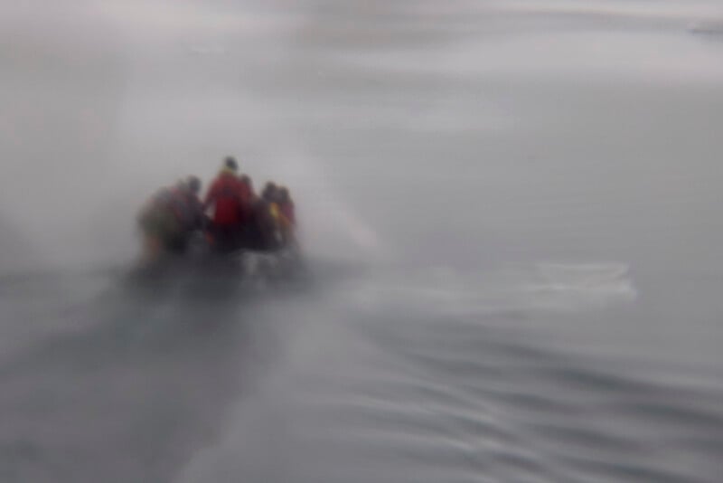 A group of people in colorful jackets ride a small inflatable boat across a misty, calm body of water. The image is blurred, creating a sense of movement and atmospheric mood.