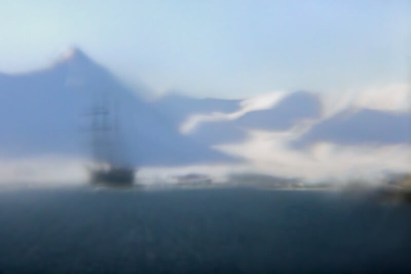 A blurry image of a sailing ship on water with snow-covered mountains in the background, shrouded in mist or fog, creating a dreamy and mysterious atmosphere.