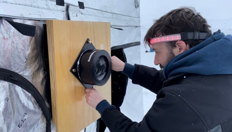 A man wearing a headband with red LED lights installs or adjusts a camera-like device mounted on a wooden panel outdoors in a snowy environment.