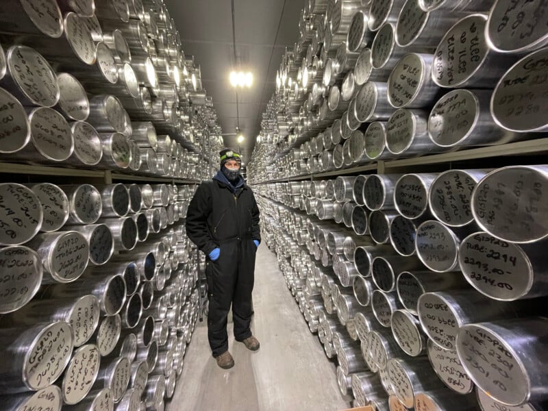 A person in winter clothing stands in a corridor between shelves stacked with rows of metal canisters, each labeled with handwritten text, inside a cold storage facility.