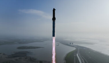 a huge silver rocket launches into a blue sky from a seaside pad