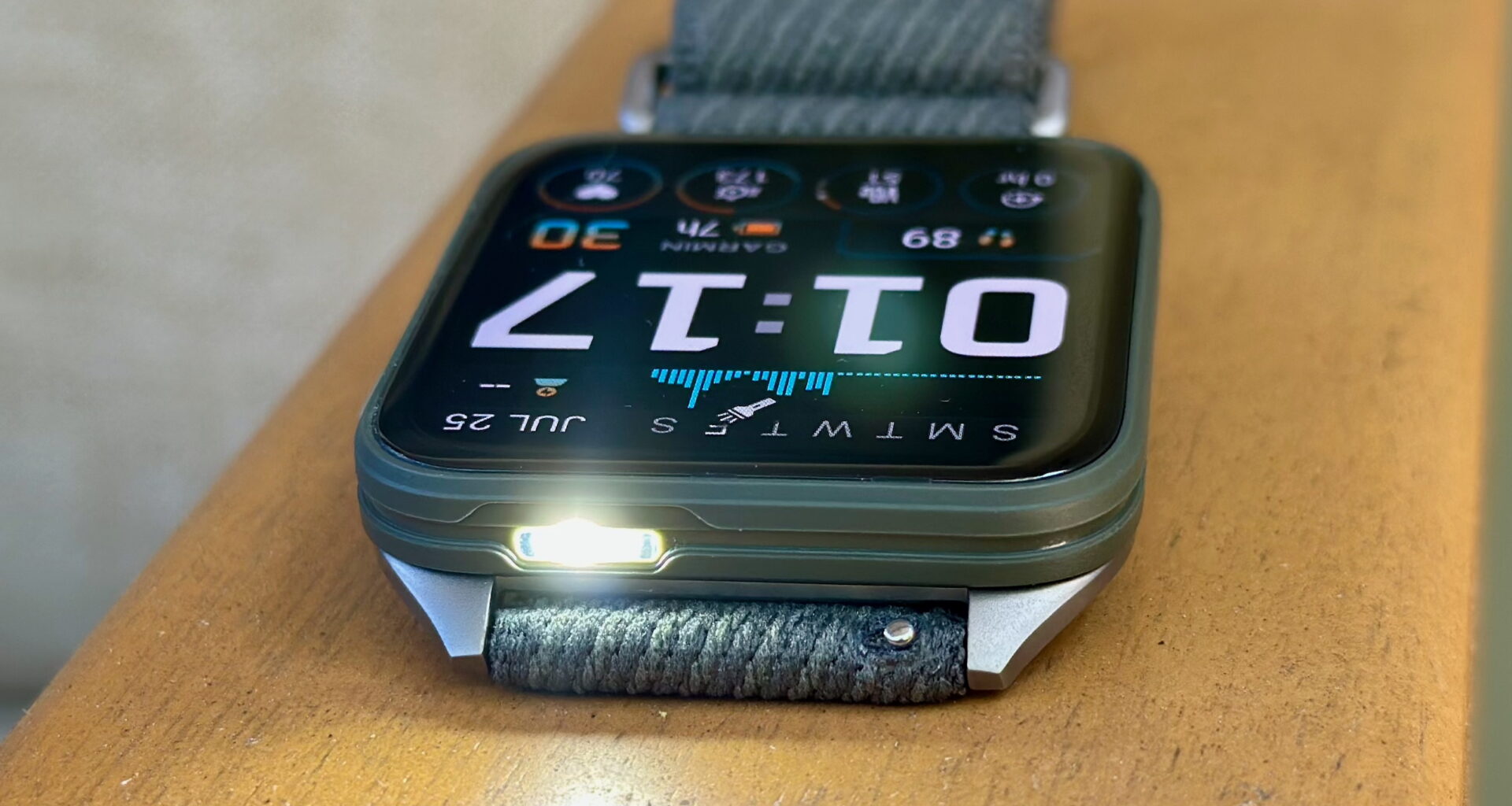 A photo of the Garmin Venu X1 sitting on an armrest, display up and facing opposite with the default watch face, the white LED flashlight on and pointing toward the camera.