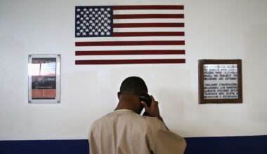 A Black man in a tan prison uniform stands in front of a black and white wall, holding a phone to his ear. A U.S. flag and two signs are on the wall. One sign reads, "Break the silence!" in English and Spanish. The second sign details that calls will subject to recording and listening.