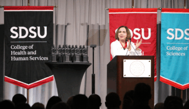 SDSU President Adela de la Torre dressed in a white blazer stands at the podium and delivers a speech during the 2025 All-University Convocation attended by hundreds
