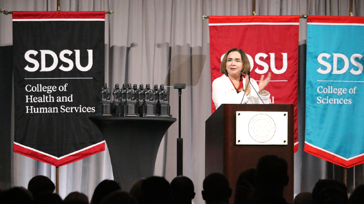 SDSU President Adela de la Torre dressed in a white blazer stands at the podium and delivers a speech during the 2025 All-University Convocation attended by hundreds