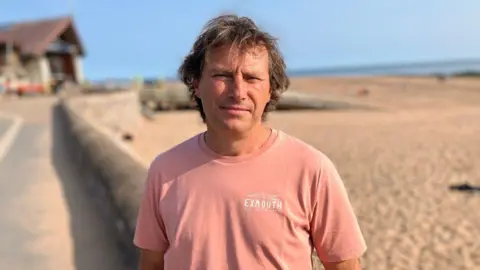 A man stands next to a beach. He is wearing a light pink top. The sand and sea is in the distance.