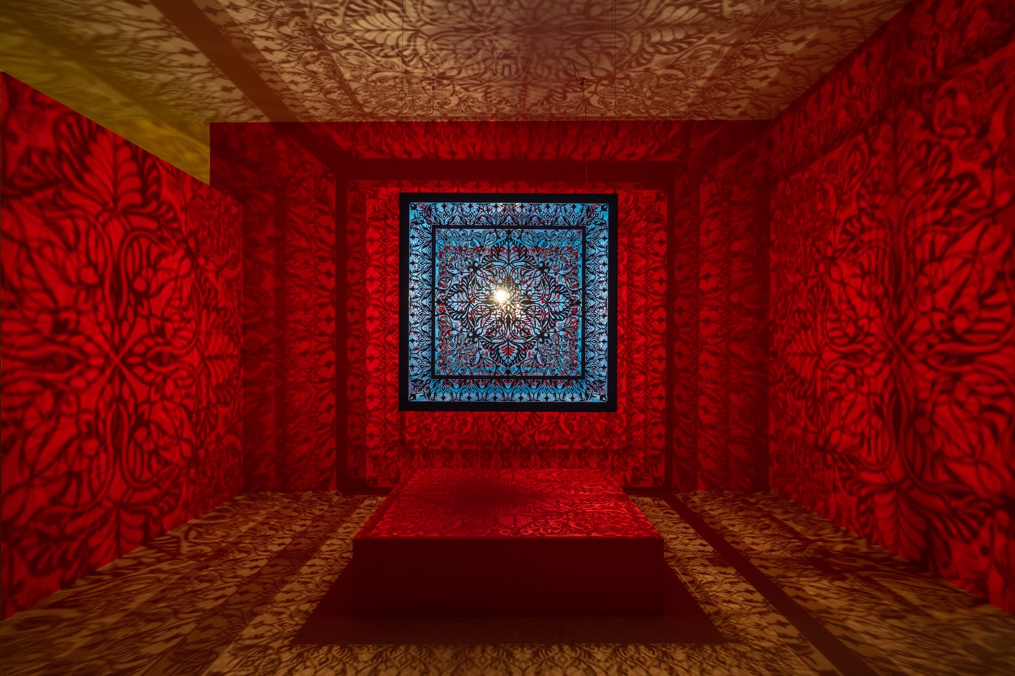 a sculptural installation in a red room, with light emitting from within a structure that casts ornate shadows onto the walls