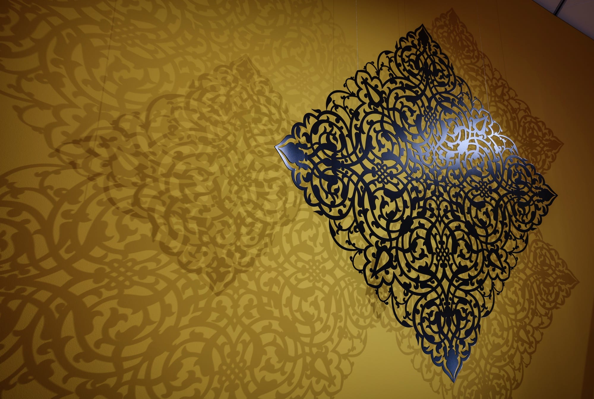 a detail of an installation of an elaborate cut-out set against a yellow wall, with light shining through to cast ornate shadows onto the wall