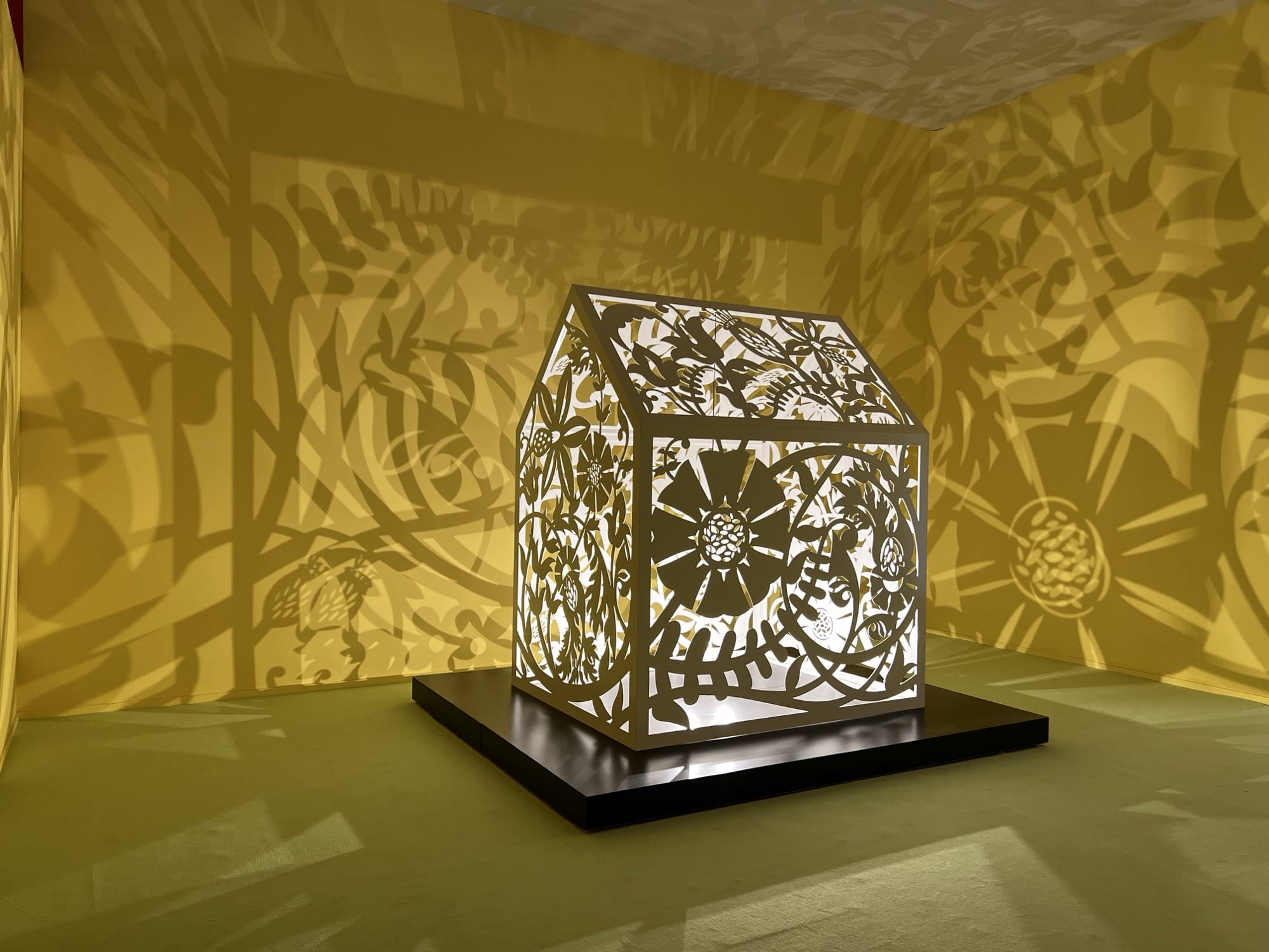 a sculptural installation in a yellow room, with light emitting from within a structure that casts ornate shadows onto the walls