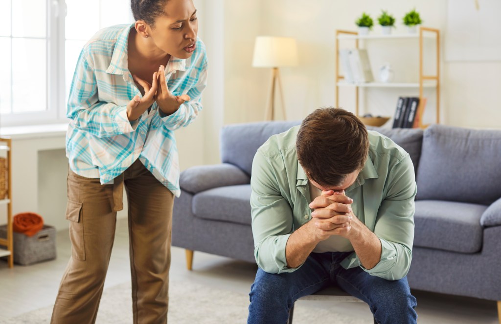Angry woman shouting at a man who is sitting with his head down.