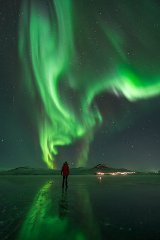 A person stands on an icy surface at night, gazing at vibrant green northern lights swirling across a star-filled sky, with distant hills and small lights on the horizon.