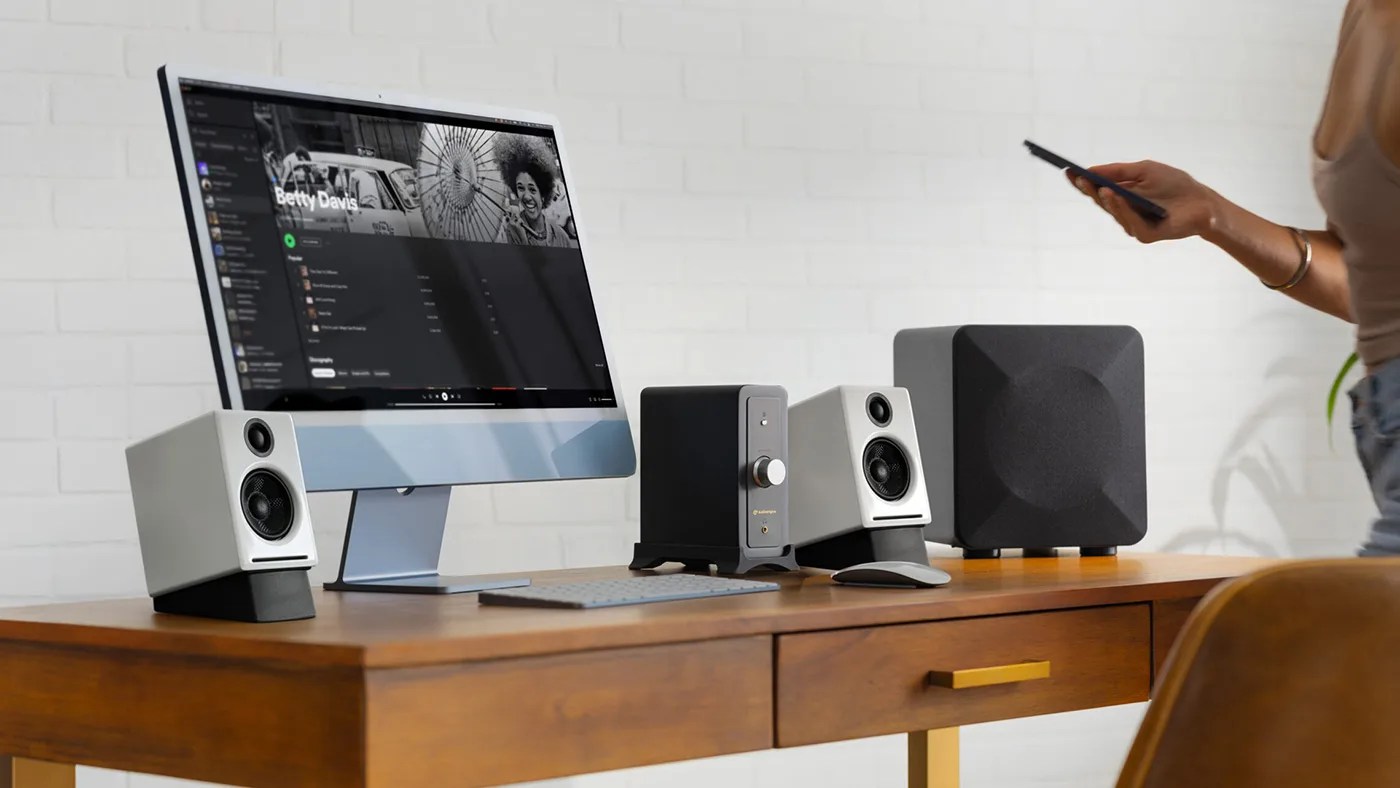 A modern wooden desk with a computer setup featuring a large monitor displaying a music playlist titled
