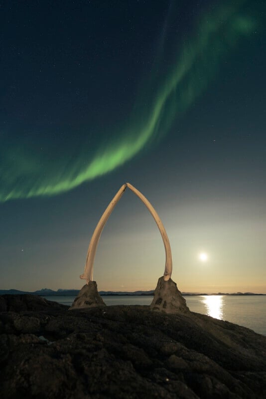 A pair of large whale bones form an arch on rocky ground by the water, illuminated by moonlight, with the green aurora borealis lighting up the night sky above.