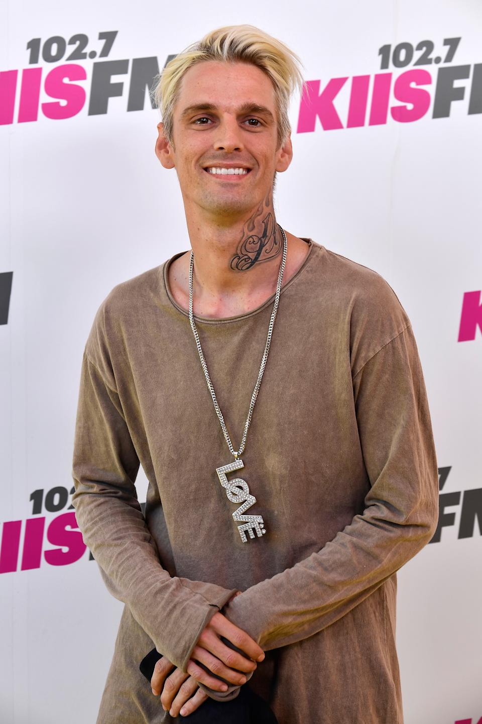 Person in casual long-sleeve top with a large "LOVE" necklace, standing in front of a backdrop with event logos