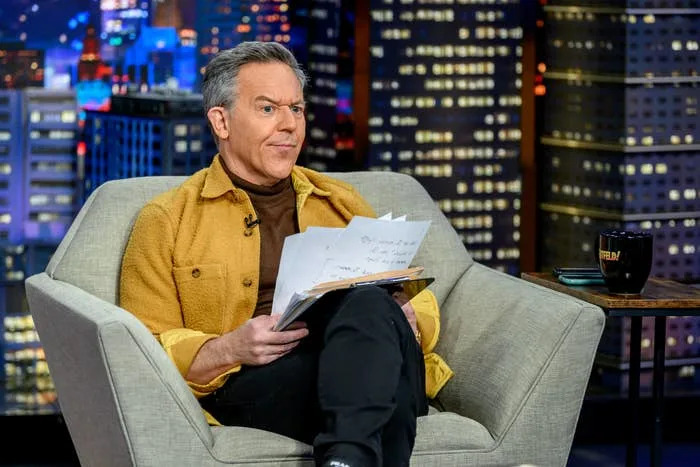 Greg Gutfeld seated, holding papers, on a talk show set with a cityscape backdrop in a casual jacket over a turtleneck sweater