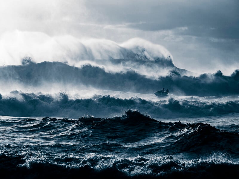 A small boat battles rough, crashing waves on a dark, stormy sea, with mist and spray rising high and large, turbulent swells in the background.