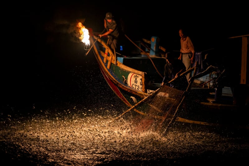 A fisherman on a boat at night holds a flaming torch over the water, attracting and illuminating a swarm of fish while another person stands nearby; splashing water and fish are visible in the darkness.