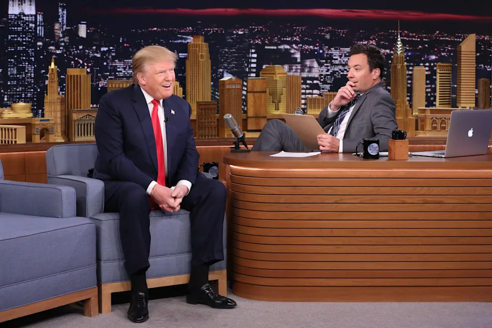 Donald Trump and Jimmy Fallon sit, talking on a late-night talk show set with a cityscape backdrop