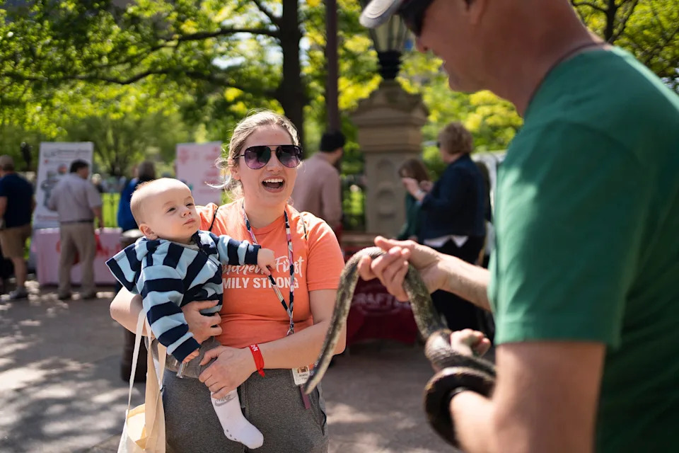 Jason Fallon, right with Ohio Department of Natural Resources, holds a gray ratsnake for Amanda Tilley, and Charlie Gould, seven months, to see, at the ODNR booth during Ohio Tourism Day on the lawn of the Ohio Statehouse in 2023.