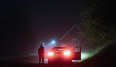 A mysterious person standing next to a car on a spooky empty road on a foggy night. Silhouetted by street lights.