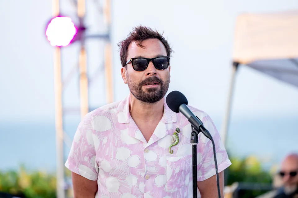 Jimmy Fallon in sunglasses and a casual pattern shirt speaks at an outdoor event with a microphone