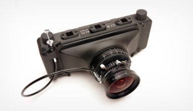 A black, compact digital camera with several labeled ports on top and a wide-angle lens attached; a cable release is connected to the camera. The camera is photographed on a white background.