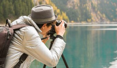 A person wearing a gray hat and jacket takes a photo with a camera on a tripod beside a calm lake, with trees and mountains in the background.