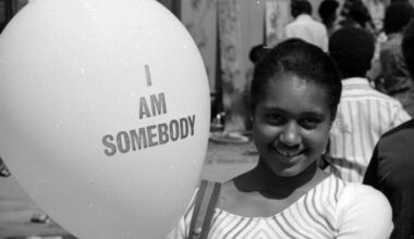 A young girl smiles while holding a balloon that reads “I AM SOMEBODY.” She stands outdoors among a crowd, wearing a striped top, with trees and people softly blurred in the background of this black-and-white photograph.