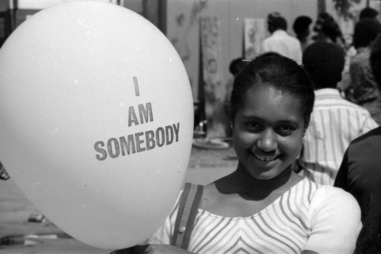A young girl smiles while holding a balloon that reads “I AM SOMEBODY.” She stands outdoors among a crowd, wearing a striped top, with trees and people softly blurred in the background of this black-and-white photograph.