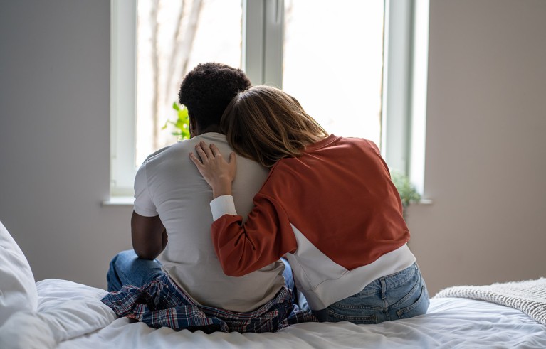 An emotional couple sit on a bed together, with a woman putting her head on a man’s shoulder for comfort and support.