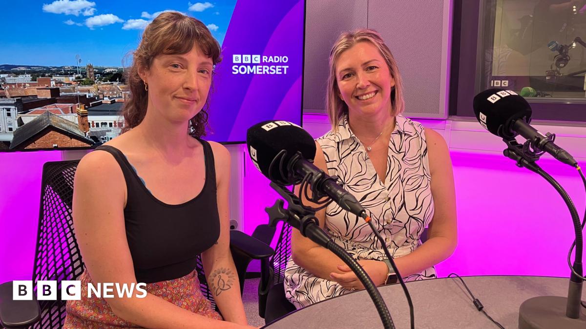 Lucy Sheppard and Aimee Peach sit next to each other in the BBC Radio Somerset studio. Lucy is wearing a black top and a skirt with a flowery pattern and Aimee is wearing a black and white dress. The studio is lit up in purple colours.