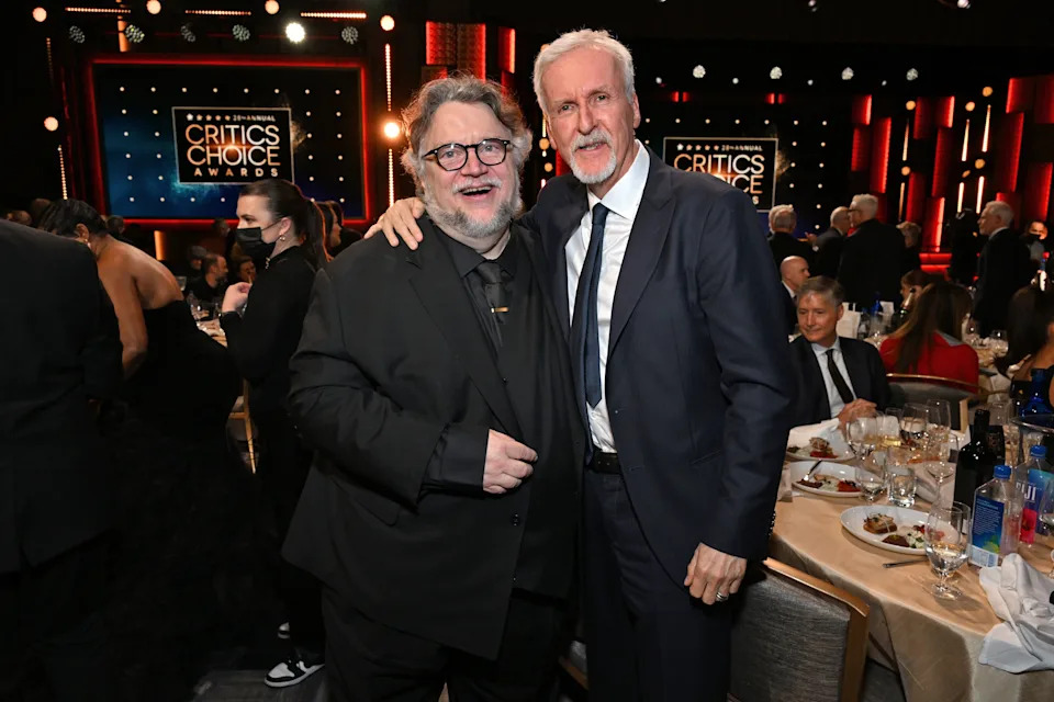 Two men in formal suits pose together at a table during the Critics Choice Awards event