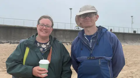 Jeannette Wilson and David Ewart standing on the beach. Jeannette has brown hair tied back, wearing glasses, a green raincoat and holding a coffee cup. David is wearing a cream hat, glasses and blue raincoat.