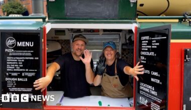 A man and woman are wearing black t-shirts and waving. They are holding open the shutters to a canal boat