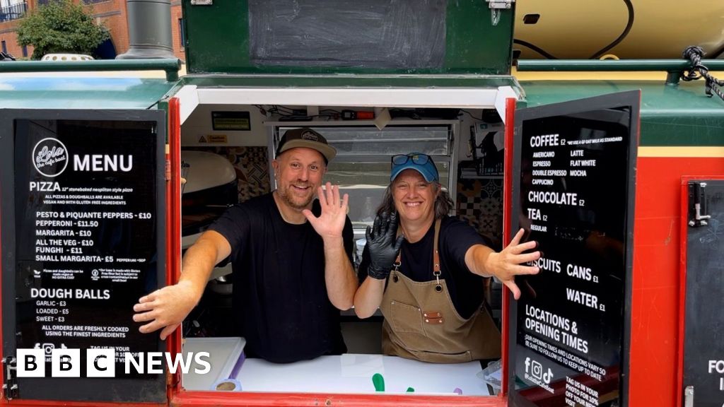 A man and woman are wearing black t-shirts and waving. They are holding open the shutters to a canal boat
