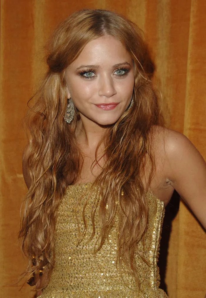 Person in glam outfit with long, wavy hair, wearing statement earrings, poses at a celebrity event