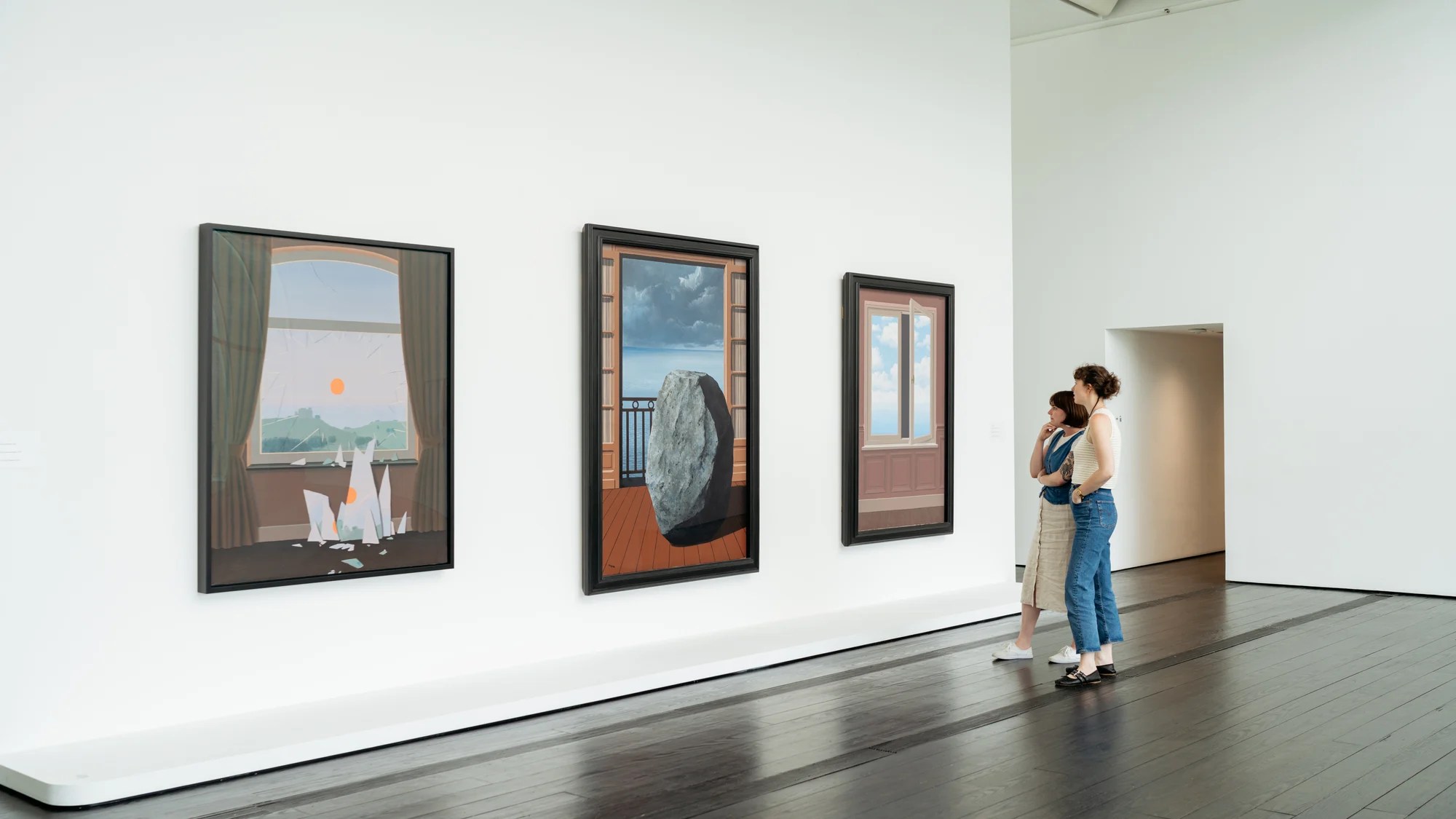 Viewers wander a gallery with three paintings hanging on the wall.