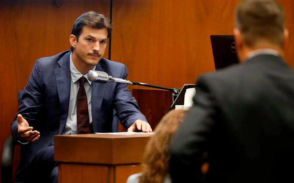 A man in a suit testifies in court, speaking into a microphone. Another person stands blurred in the foreground