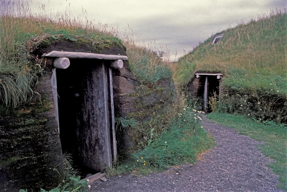 Earth-covered houses with grass roofs and wooden doors, blending into the landscape. Gravel path leads to entrances. Overcast sky in the background