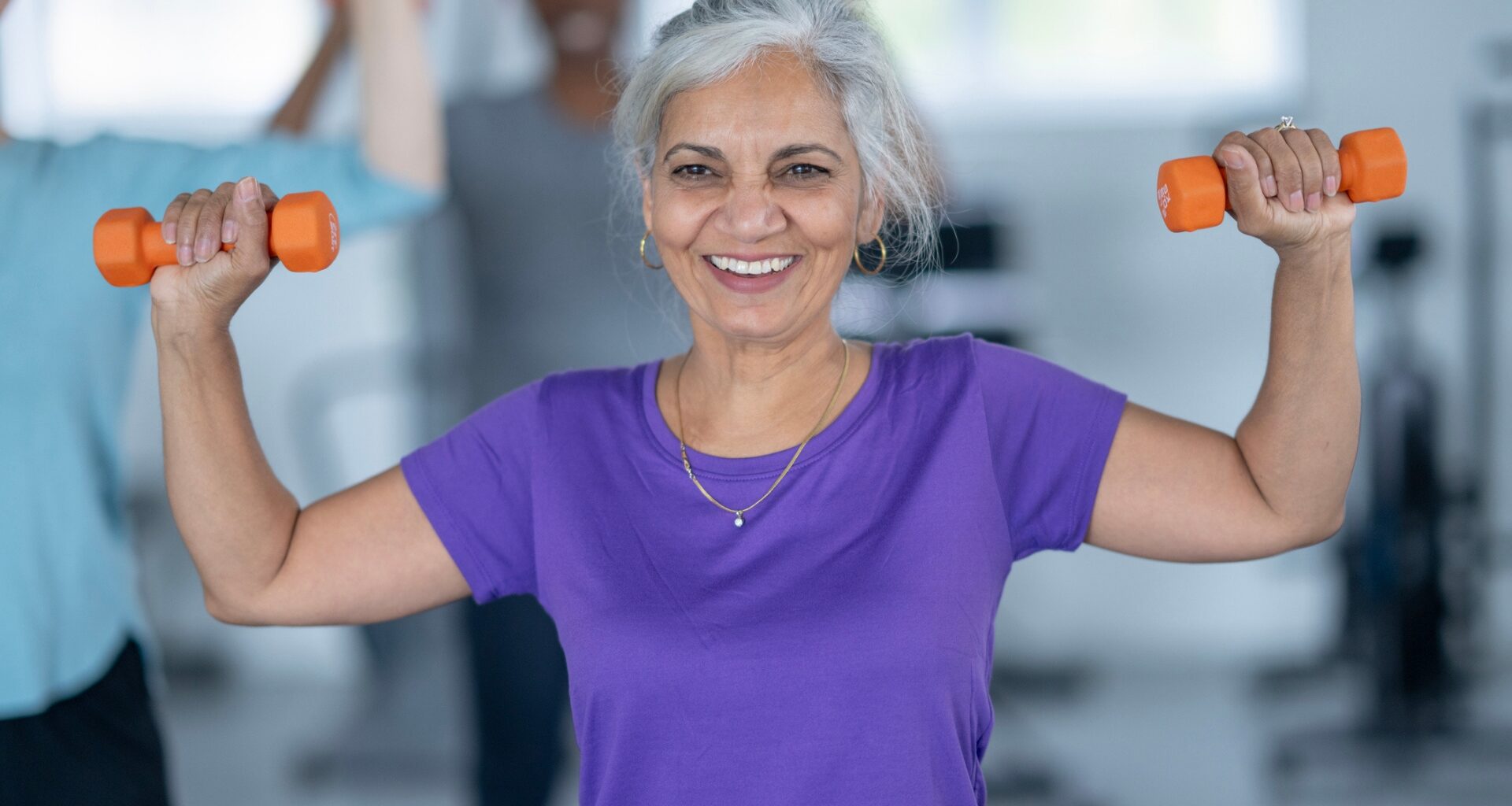 woman wearing a purple tshirt facing the camera, smiling and holding a small orange dumbbell in each hand at shoulder height half way through an overhead press.