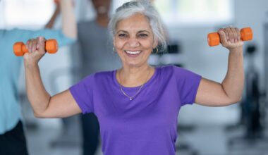 woman wearing a purple tshirt facing the camera, smiling and holding a small orange dumbbell in each hand at shoulder height half way through an overhead press.