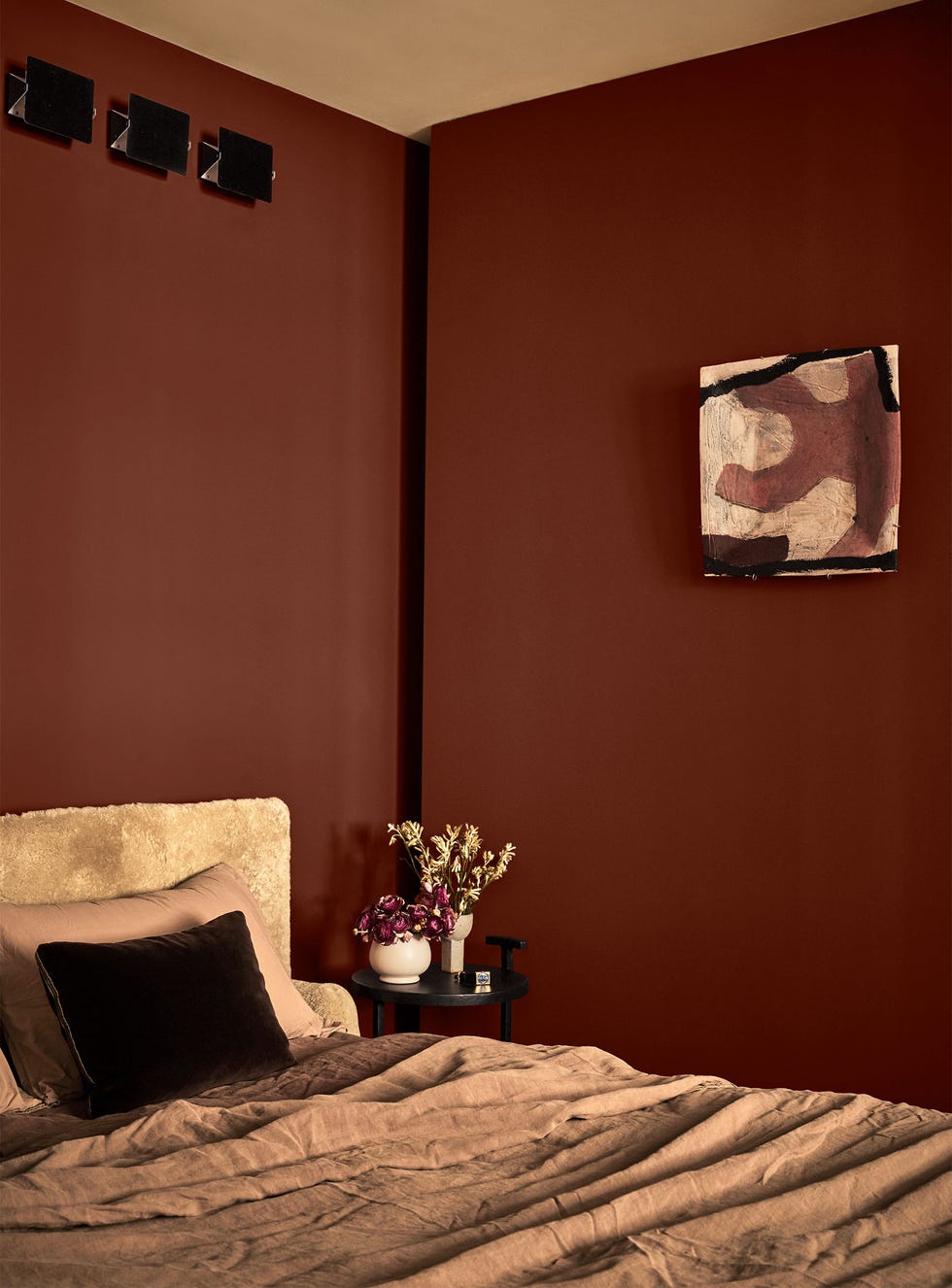 in a brown painted bedroom, a bed's headboard is covered in a light brown shearling, bed covering is in a medium brown linen, a small round night table has a plant and a vase with flowers, a small artwork on wall