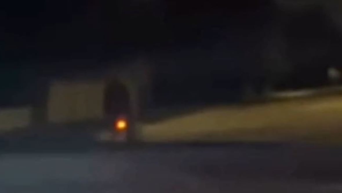 Someone riding their bike on a suburban street at nighttime is seen through a vehicle windshield. The bike's rear reflector shines bright from the vehicle's headlights.