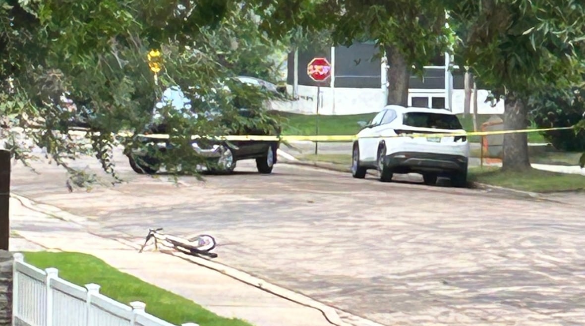 A bicycle lies on the curb of a suburban street. Just north of it, yellow police tape blocks off the street at a nearby intersection, and a black SUV is parked in front of the tape.